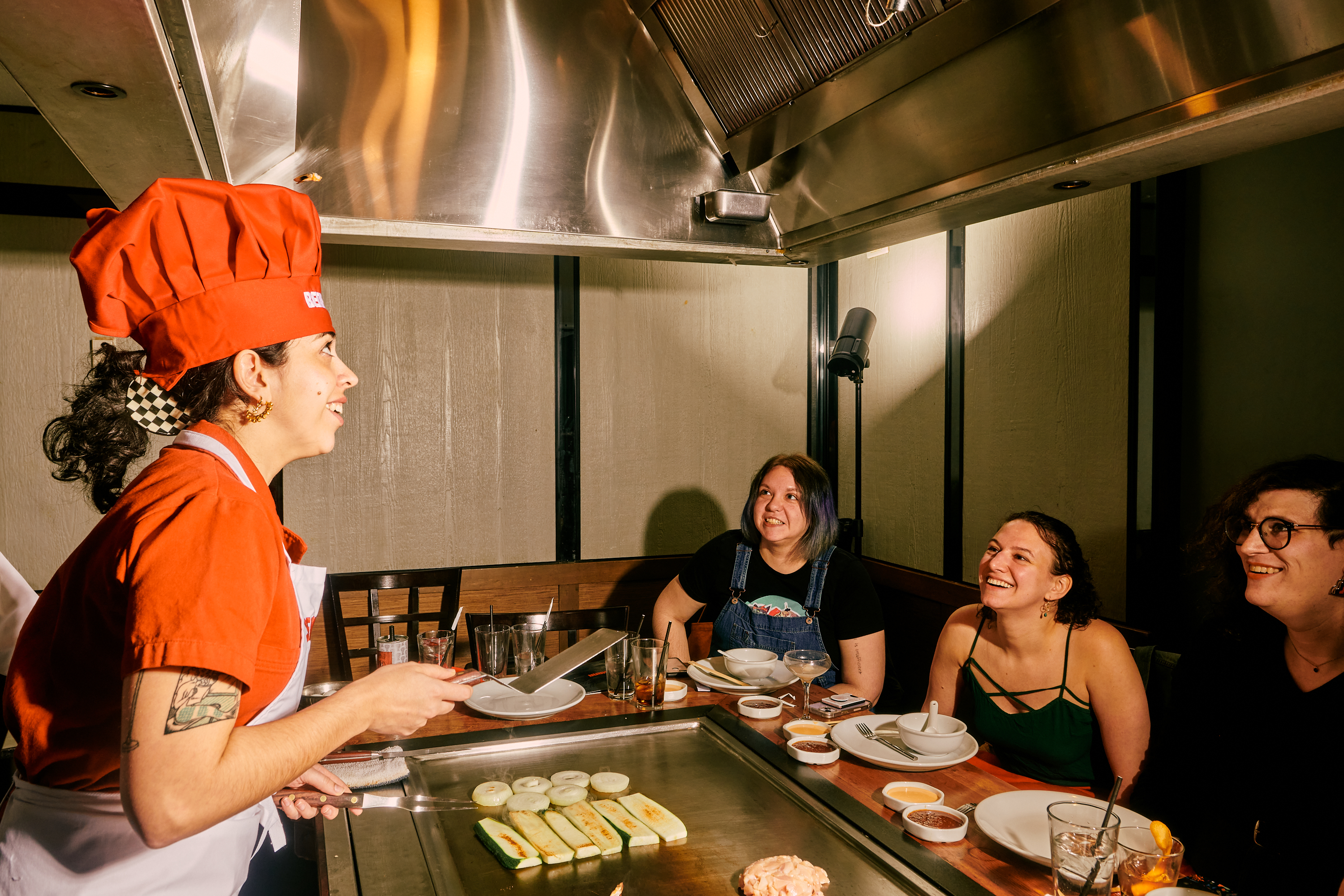 Jaya, standing behind Benihana’s grill, flips a shrimp tail into her chef’s hat as her friends look on, smiling.
