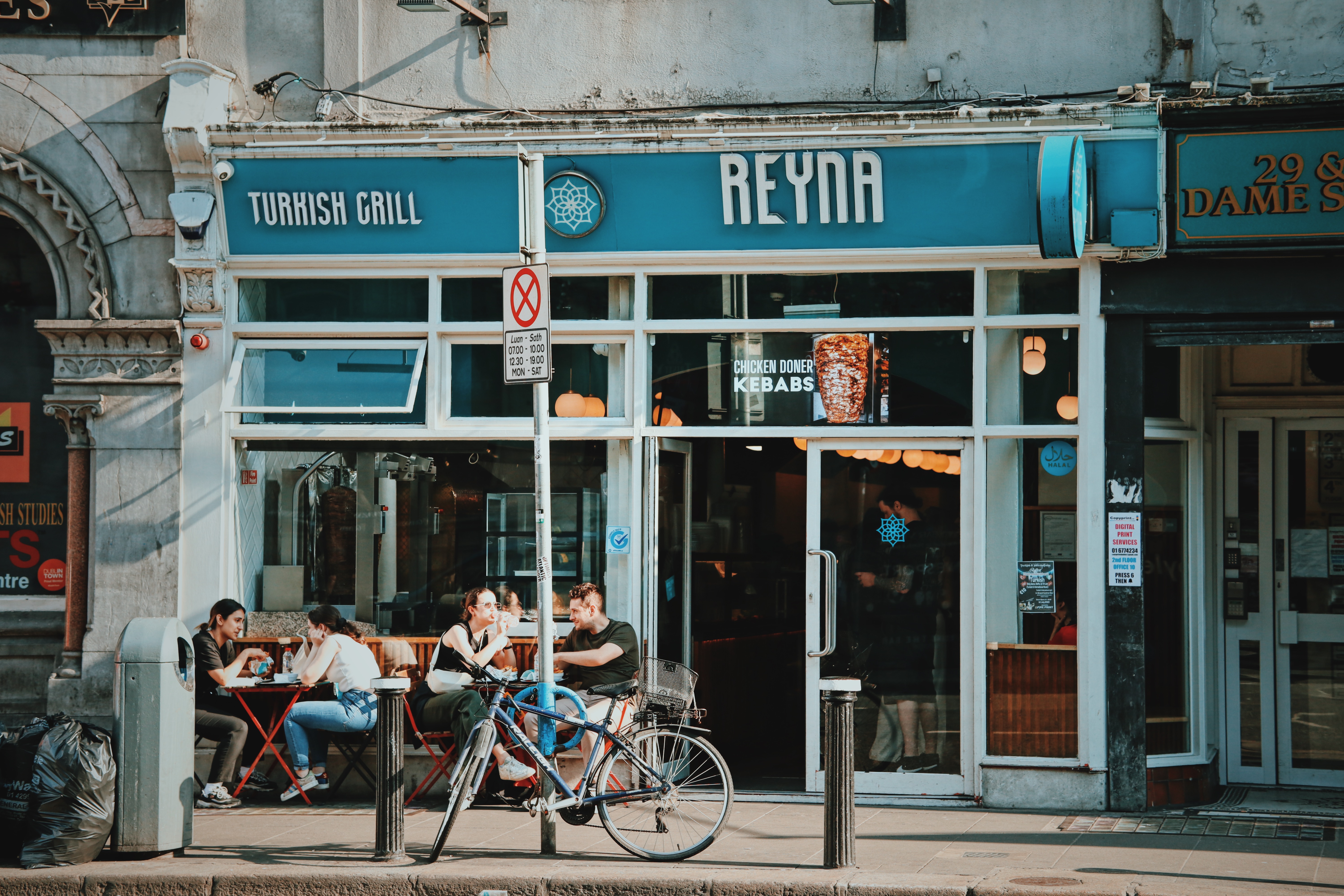 Customers sit at tables outside a restaurant with signs advertising Reyna and Turkish Grill.