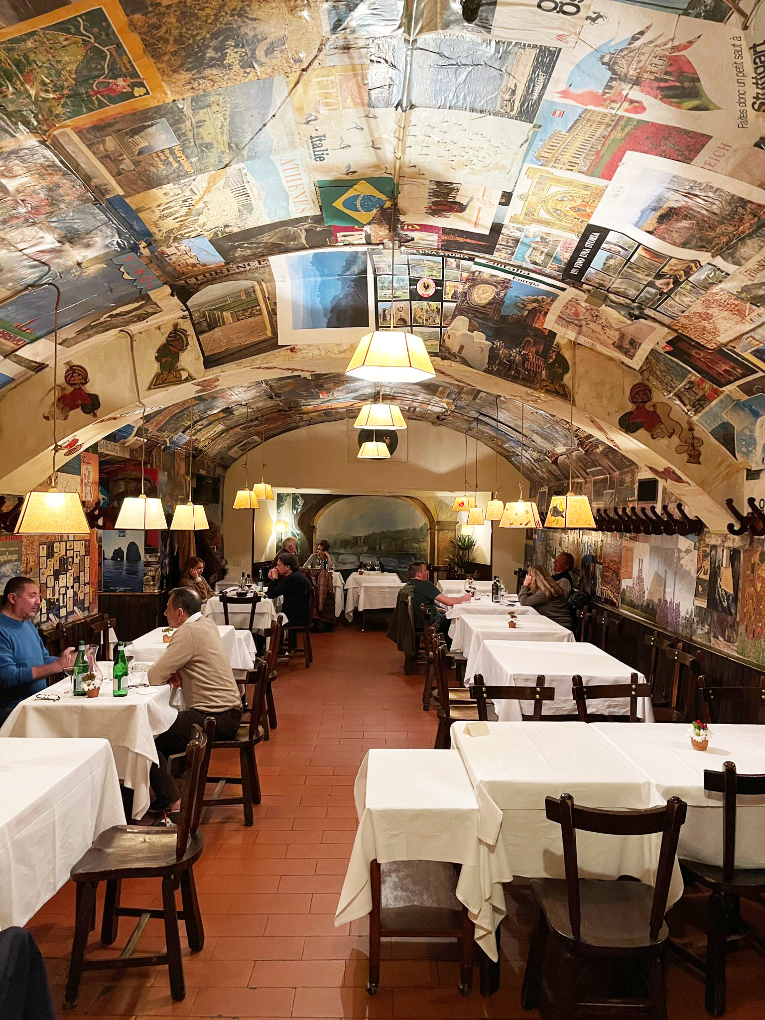 A restaurant interior where the arched, cave-like ceiling and walls are covered with old posters, diners sit at white tablecloth-covered tables beneath pendant lights.